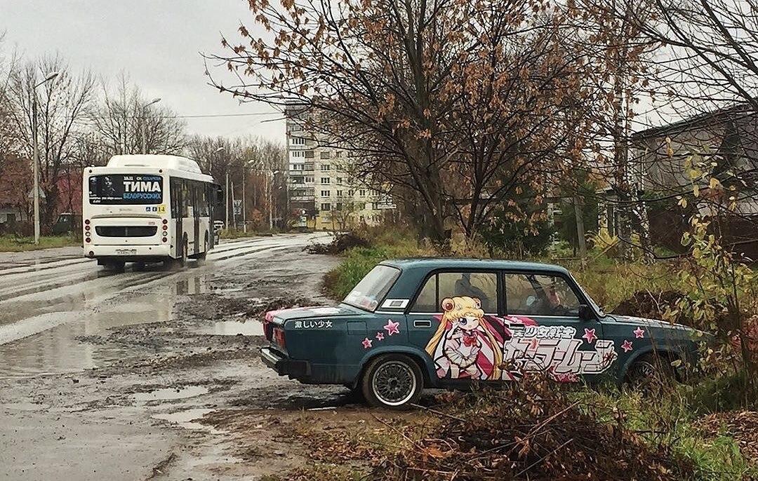 A picture of a rainy day, with a green car in the foreground. It's an older model, but sports an anime decal across the entire side.