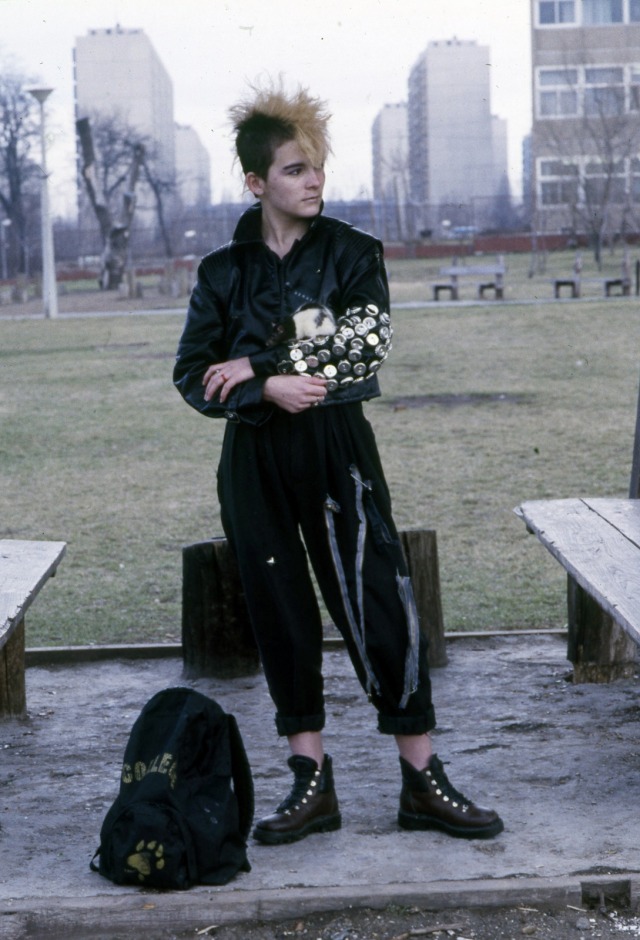 A young androgenous person, with blond hair spiked in a messy mohawk and black loose clothes. Their left sleeve is completely covered in silver pins, making it look like they are holding a bouquet of silver flowers.