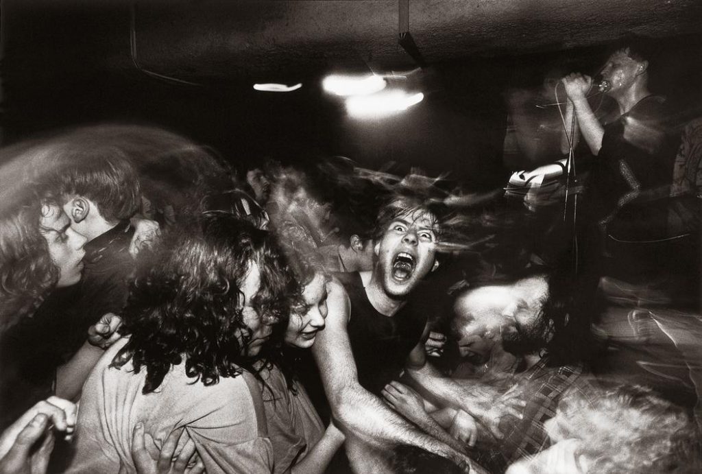 A dark, poorly lit concert full of young people and smoke. A singer on stage with his microphone is barely visible to the right. The centerpiece of the photo is a young man, who appears to be screaming at the camera. 