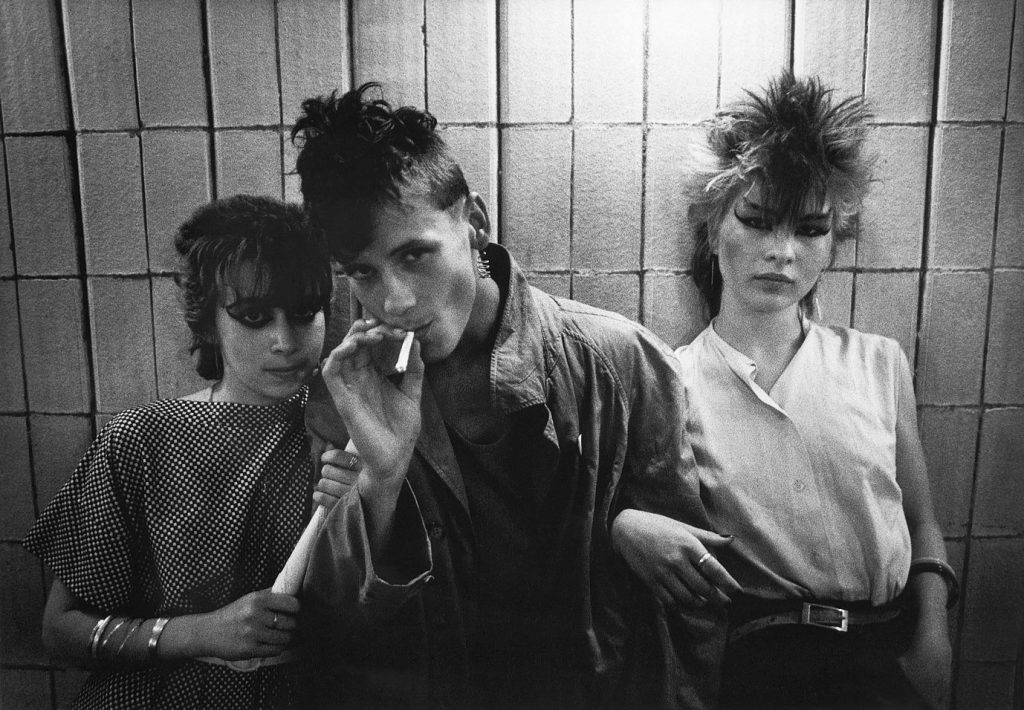 Three young people, two women in vivid black eyeliner and a man with intense eyes stand against the wall. The man is smoking a cigarette, and appears to be making a statement by looking directly into the camera.